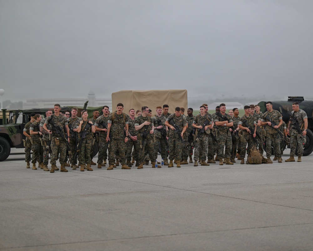 A large group of military personnel in woodland MARPAT camouflage uniforms are assembled on what appears to be a flight line or tarmac at a military airfield. The personnel are carrying rifles and wearing combat gear, suggesting a deployment, exercise, or formation event. The overcast, hazy weather conditions and airfield infrastructure are visible in the background.