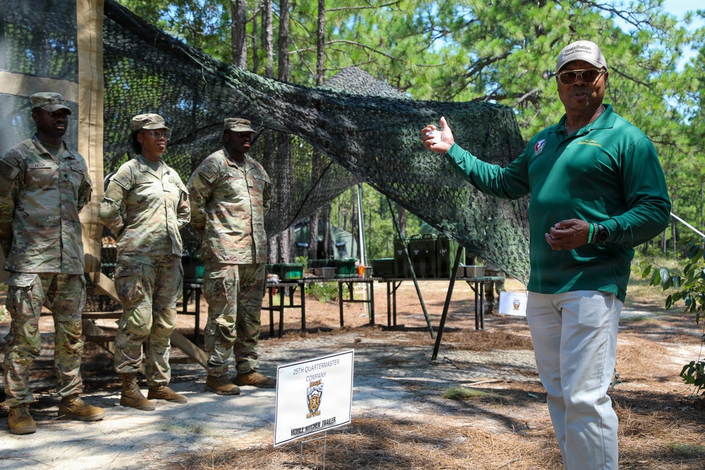 A civilian in a green polo shirt appears to be briefing or addressing three soldiers in Army Combat Uniforms (ACUs) at an outdoor military field site. A sign in the foreground identifies this as the '29th Quartermaster Company Mobile Kitchen Trailer' setup. The setting includes camouflage netting, field tables with equipment, and a wooded environment consistent with a military training area.