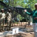 A civilian in a green polo shirt appears to be briefing or addressing three soldiers in Army Combat Uniforms (ACUs) at an outdoor military field site. A sign in the foreground identifies this as the '29th Quartermaster Company Mobile Kitchen Trailer' setup. The setting includes camouflage netting, field tables with equipment, and a wooded environment consistent with a military training area.