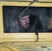 A soldier wearing a military helmet is visible through the windshield of a desert-tan colored military Humvee (HMMWV). The vehicle's exterior is painted in a sand/tan color typical of desert operations. A notepad and what appears to be a red pen or marker are visible on the dashboard inside the vehicle.