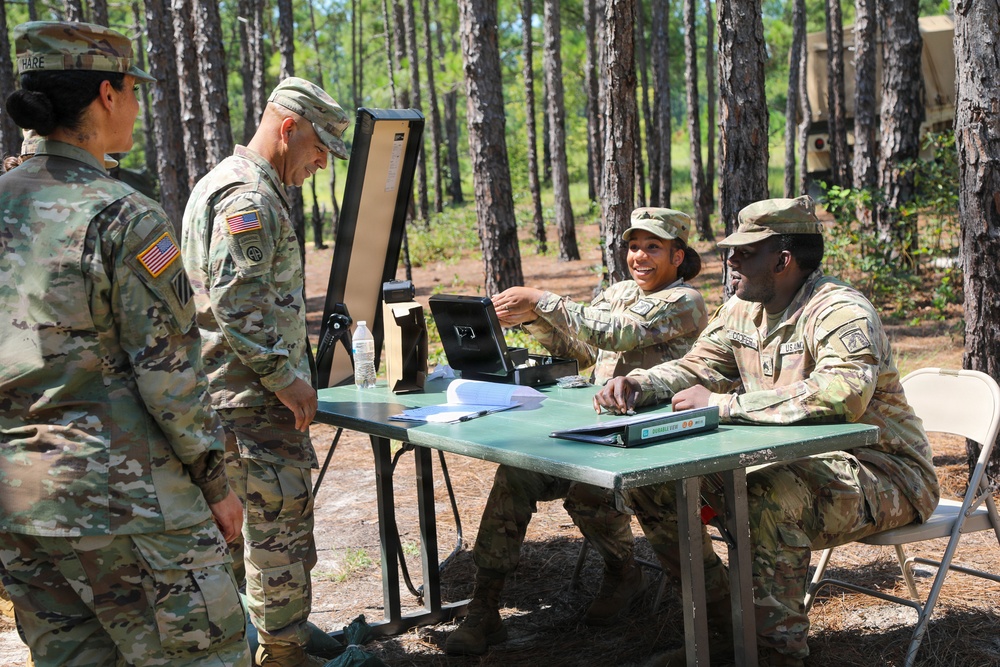 Four U.S. Army soldiers in OCP (Operational Camouflage Pattern) uniforms are gathered around a folding table in a wooded outdoor setting. Two soldiers are seated behind the table operating what appears to be a tablet or electronic device on a stand, while two others stand on the opposite side. The setting suggests a field exercise, checkpoint, or administrative station.