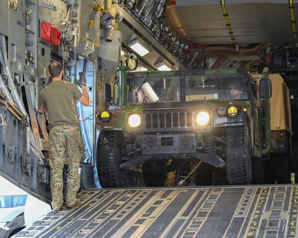 A military HMMWV (Humvee) is being loaded or unloaded from a large military transport aircraft, likely a C-17 Globemaster III, as indicated by the distinctive cargo bay interior. At least two personnel are visible inside the Humvee while a third soldier in MultiCam/OCP uniform guides the vehicle from the side. The vehicle's headlights are illuminated and the surface appears wet, suggesting recent rain or washing.
