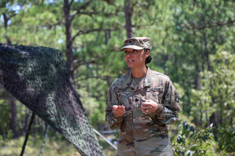 A U.S. Army officer wearing Operational Camouflage Pattern (OCP) uniform is standing outdoors in a wooded area, appearing to speak or brief someone. The name tape reads 'HARE' and a gold oak leaf insignia is visible on the chest, indicating the rank of Major. A camouflage netting structure is visible in the background among pine trees.