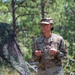 A U.S. Army officer wearing Operational Camouflage Pattern (OCP) uniform is standing outdoors in a wooded area, appearing to speak or brief someone. The name tape reads 'HARE' and a gold oak leaf insignia is visible on the chest, indicating the rank of Major. A camouflage netting structure is visible in the background among pine trees.