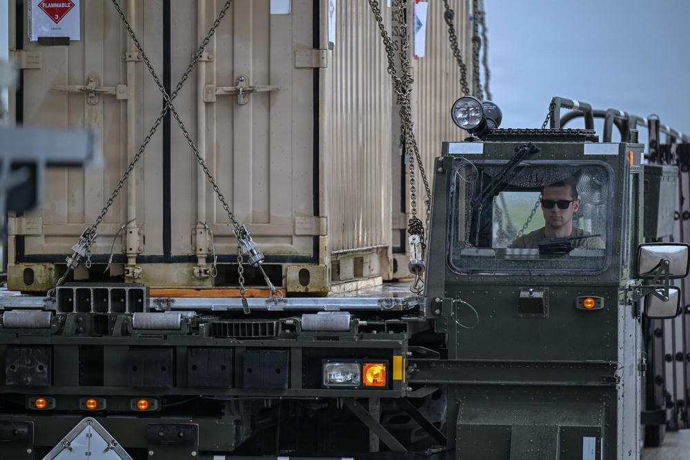 A military vehicle operator in sunglasses is seated in the cab of an olive-drab military truck, maneuvering or positioning large tan military containers secured with chains on flatbed trailers. The containers bear a red 'FLAMMABLE' hazard diamond placard, indicating hazardous cargo. The scene appears to be a military logistics or loading operation, likely at an airfield or staging area.