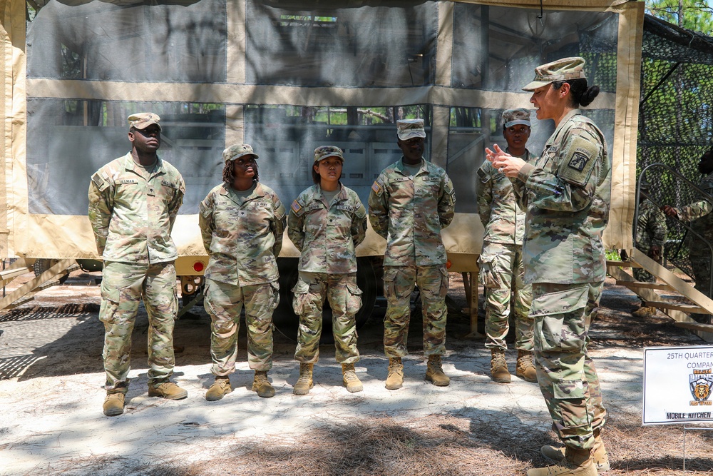 A U.S. Army soldier, likely a non-commissioned officer or officer, is briefing or instructing a group of four soldiers in front of a field mobile kitchen setup. A sign visible in the lower right reads '25th Quartermaster Company' and 'Mobile Kitchen.' The setting appears to be an outdoor field training exercise in a wooded area.