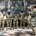 A U.S. Army soldier, likely a non-commissioned officer or officer, is briefing or instructing a group of four soldiers in front of a field mobile kitchen setup. A sign visible in the lower right reads '25th Quartermaster Company' and 'Mobile Kitchen.' The setting appears to be an outdoor field training exercise in a wooded area.