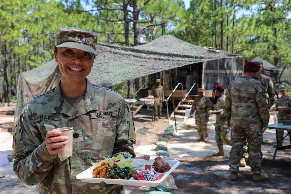 A U.S. Army soldier identified by name tape as 'HARE' smiles while holding a food tray and beverage cup at what appears to be a field dining facility. The setting is an outdoor military camp in a forested area with camouflage netting overhead. Several other soldiers in OCP uniforms are visible in the background near a field kitchen structure.