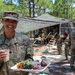 A U.S. Army soldier identified by name tape as 'HARE' smiles while holding a food tray and beverage cup at what appears to be a field dining facility. The setting is an outdoor military camp in a forested area with camouflage netting overhead. Several other soldiers in OCP uniforms are visible in the background near a field kitchen structure.