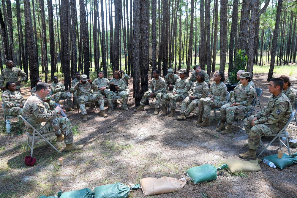A group of approximately 20 military personnel in US Army OCP (Operational Camouflage Pattern) uniforms are seated in a circular formation on folding chairs in a pine forest clearing. One individual on the left appears to be leading a discussion or briefing, addressing the group. The setting suggests an outdoor training session, leadership development meeting, or after-action review.