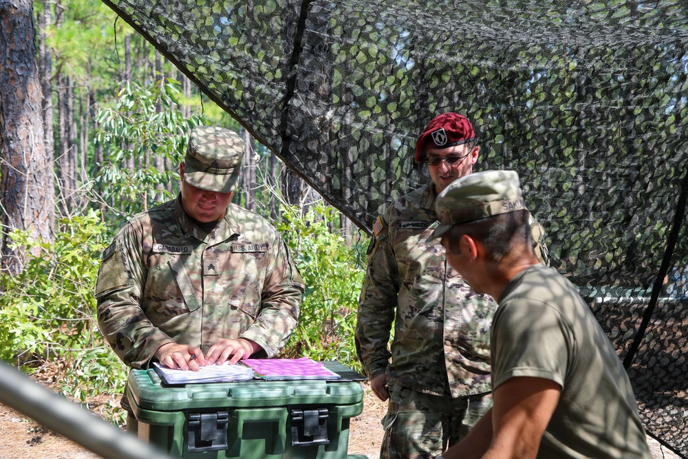 Three U.S. Army soldiers are gathered under a camouflage netting in a wooded outdoor setting. One soldier, identified by name tape as 'CARABALLO' with a Sergeant rank insignia, is reviewing documents placed on a green military storage case. A second soldier wearing a maroon/red beret is observing, while a third soldier in a patrol cap is partially visible in the foreground.
