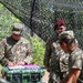 Three U.S. Army soldiers are gathered under a camouflage netting in a wooded outdoor setting. One soldier, identified by name tape as 'CARABALLO' with a Sergeant rank insignia, is reviewing documents placed on a green military storage case. A second soldier wearing a maroon/red beret is observing, while a third soldier in a patrol cap is partially visible in the foreground.