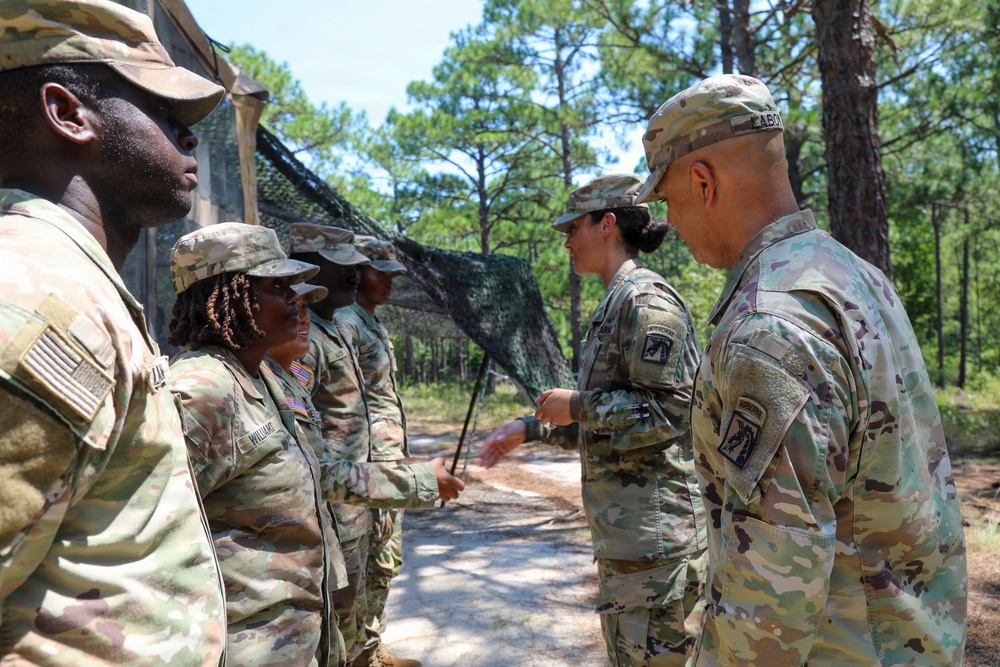 A group of U.S. Army soldiers in Operational Camouflage Pattern (OCP) uniforms are gathered outdoors in a wooded area, appearing to be engaged in a formal interaction or ceremony. A senior soldier on the right is facing and interacting with soldiers in a line, possibly conducting an inspection or presenting coins/awards. The setting appears to be a field training environment with camouflage netting visible in the background.