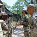 A group of U.S. Army soldiers in Operational Camouflage Pattern (OCP) uniforms are gathered outdoors in a wooded area, appearing to be engaged in a formal interaction or ceremony. A senior soldier on the right is facing and interacting with soldiers in a line, possibly conducting an inspection or presenting coins/awards. The setting appears to be a field training environment with camouflage netting visible in the background.