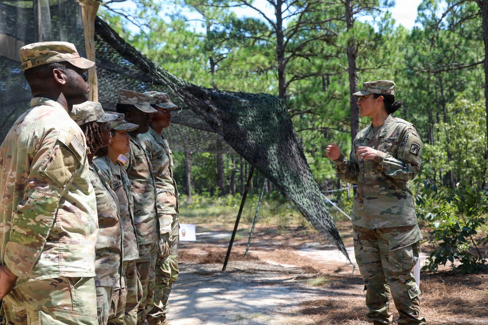 A group of U.S. Army soldiers in MultiCam/OCP camouflage uniforms are gathered in a wooded outdoor training area. One soldier on the right appears to be briefing or instructing the others who are standing in a line. A camouflage netting structure is erected in the background, suggesting a field training exercise environment.