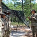 A group of U.S. Army soldiers in MultiCam/OCP camouflage uniforms are gathered in a wooded outdoor training area. One soldier on the right appears to be briefing or instructing the others who are standing in a line. A camouflage netting structure is erected in the background, suggesting a field training exercise environment.