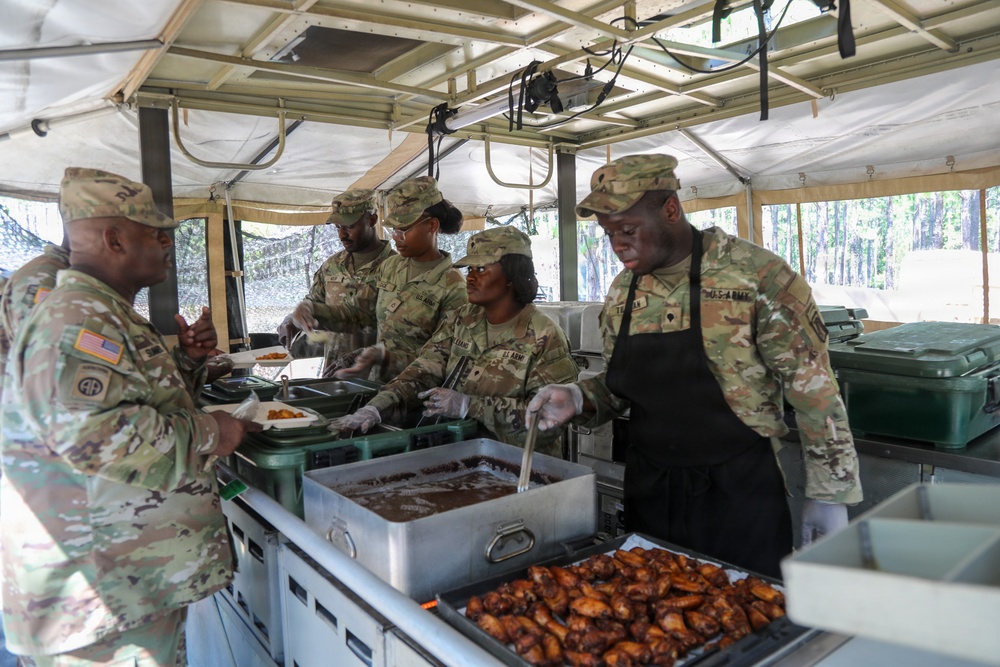 U.S. Army soldiers are operating a field kitchen under a military tent structure, serving hot meals in what appears to be a field training environment. Several soldiers wearing OCP (Operational Camouflage Pattern) uniforms and black aprons are serving food from large metal trays to fellow soldiers. The setting includes standard military field kitchen equipment and appears to be located in a wooded area.