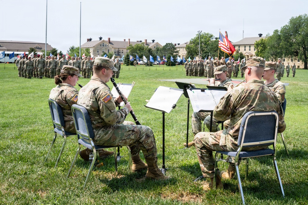 A small ensemble of U.S. Army soldiers in OCP (Operational Camouflage Pattern) uniforms performs music outdoors on a military installation parade field. They are seated in folding chairs with music stands, playing woodwind and brass instruments during what appears to be a formal military ceremony. In the background, large formations of soldiers stand at attention alongside multiple unit guidons and flags, including the American flag.