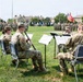 A small ensemble of U.S. Army soldiers in OCP (Operational Camouflage Pattern) uniforms performs music outdoors on a military installation parade field. They are seated in folding chairs with music stands, playing woodwind and brass instruments during what appears to be a formal military ceremony. In the background, large formations of soldiers stand at attention alongside multiple unit guidons and flags, including the American flag.