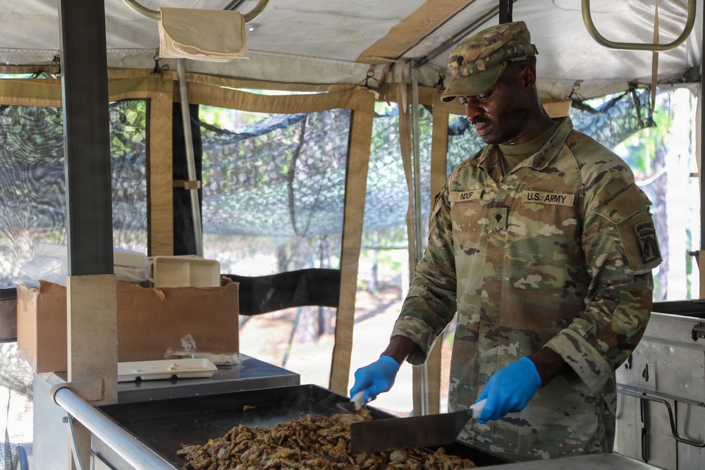 A U.S. Army soldier is preparing food on a field cooking griddle under a military field kitchen tent. The soldier, identified by the name tape 'NOOP' and 'U.S. ARMY' tape on their OCP (Operational Camouflage Pattern) uniform, is using a spatula to cook what appears to be ground meat or similar food. The setting appears to be a forward operating base or field training exercise with camouflage netting visible in the background.