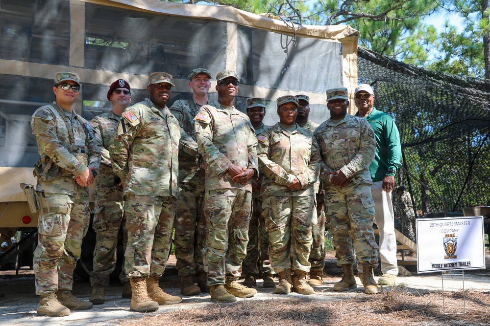 A group of approximately ten individuals, mostly in U.S. Army OCP (Operational Camouflage Pattern) uniforms, pose for a group photo in front of a Mobile Kitchen Trailer belonging to the 29th Quartermaster Company. One individual on the far right is wearing civilian clothing (green shirt and khaki pants), suggesting a civilian visitor or dignitary. The setting appears to be an outdoor military field exercise or demonstration area with pine trees in the background.