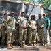 A group of approximately ten individuals, mostly in U.S. Army OCP (Operational Camouflage Pattern) uniforms, pose for a group photo in front of a Mobile Kitchen Trailer belonging to the 29th Quartermaster Company. One individual on the far right is wearing civilian clothing (green shirt and khaki pants), suggesting a civilian visitor or dignitary. The setting appears to be an outdoor military field exercise or demonstration area with pine trees in the background.