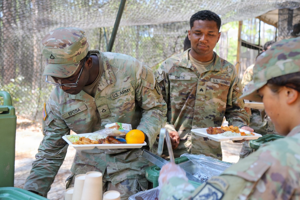 U.S. Army soldiers are going through a field chow line at an outdoor military dining facility. Soldiers identified by name tapes 'EFOSA' and 'SMITH' are receiving food on trays in what appears to be a field training environment. The setting includes camouflage netting overhead and portable military field kitchen equipment.