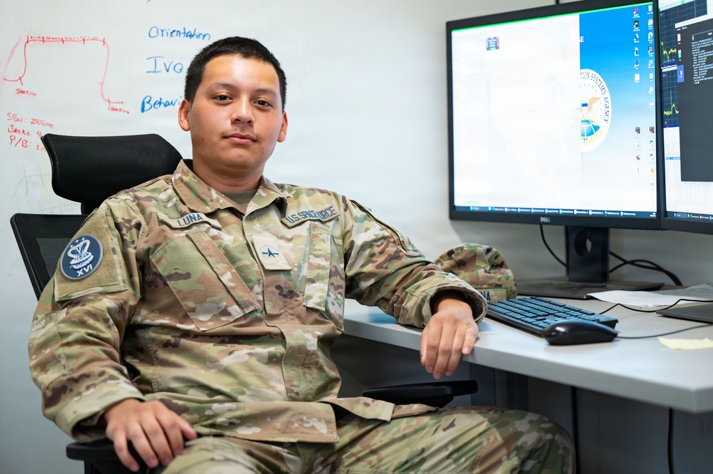 A U.S. Space Force member with the name tag 'Luna' and rank insignia of a 2nd Lieutenant is seated at a workstation with dual Dell monitors. The individual is wearing OCP (Operational Camouflage Pattern) uniform with a U.S. Space Force chest tape and a XVI unit patch on the left sleeve. The setting appears to be an office or operations center environment.