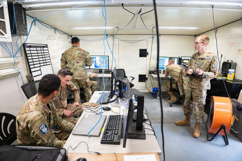 Military personnel in OCP (Operational Camouflage Pattern) uniforms are working inside what appears to be a tactical operations center or mobile command post. Multiple soldiers are engaged with computer workstations, reviewing data on monitors, and consulting with each other. The environment is a confined, utilitarian space with visible cabling infrastructure, whiteboards, and cooling equipment.