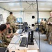 Military personnel in OCP (Operational Camouflage Pattern) uniforms are working inside what appears to be a tactical operations center or mobile command post. Multiple soldiers are engaged with computer workstations, reviewing data on monitors, and consulting with each other. The environment is a confined, utilitarian space with visible cabling infrastructure, whiteboards, and cooling equipment.