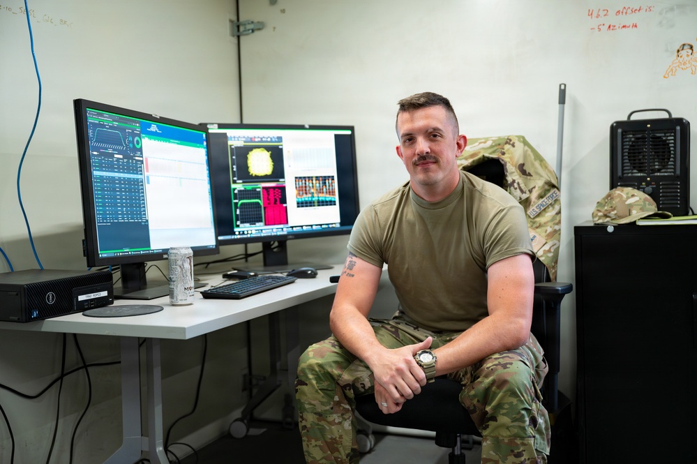 A U.S. Air Force service member in OCP (Operational Camouflage Pattern) uniform sits at a workstation with dual monitors displaying what appears to be radar or sensor data interfaces. The setting appears to be a military operations or technical workspace, with notes written on a whiteboard wall behind him. The individual is posing for what appears to be an official or editorial photograph.