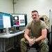 A U.S. Air Force service member in OCP (Operational Camouflage Pattern) uniform sits at a workstation with dual monitors displaying what appears to be radar or sensor data interfaces. The setting appears to be a military operations or technical workspace, with notes written on a whiteboard wall behind him. The individual is posing for what appears to be an official or editorial photograph.