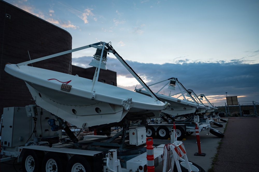 The image shows a row of large mobile satellite dish antennas mounted on wheeled trailers, parked in an outdoor lot adjacent to a dark brick building at dusk. The dishes are tilted at an angle and appear to be in a stowed or transport position. Orange traffic cones and safety barriers are visible nearby, suggesting a staging or operational area.
