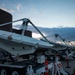 The image shows a row of large mobile satellite dish antennas mounted on wheeled trailers, parked in an outdoor lot adjacent to a dark brick building at dusk. The dishes are tilted at an angle and appear to be in a stowed or transport position. Orange traffic cones and safety barriers are visible nearby, suggesting a staging or operational area.
