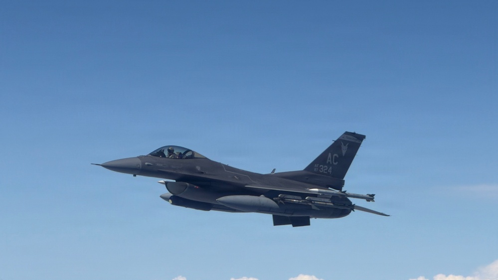 A single-seat F-16 Fighting Falcon jet fighter is photographed in flight against a clear blue sky. The aircraft bears tail code 'AC' and serial number '324', indicating it belongs to the New Jersey Air National Guard's 177th Fighter Wing based at Atlantic City. The aircraft is carrying external stores including what appear to be fuel tanks and air-to-air missiles.