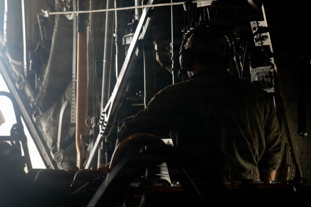 A silhouetted military crew member wearing aviation headset/helmet is seated inside what appears to be a large military transport aircraft, likely near an open cargo ramp or door. The interior structural elements of the aircraft are visible, with bright light entering from the opening. The scene suggests an in-flight operation, possibly monitoring a cargo drop or airdrop procedure.