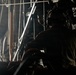 A silhouetted military crew member wearing aviation headset/helmet is seated inside what appears to be a large military transport aircraft, likely near an open cargo ramp or door. The interior structural elements of the aircraft are visible, with bright light entering from the opening. The scene suggests an in-flight operation, possibly monitoring a cargo drop or airdrop procedure.