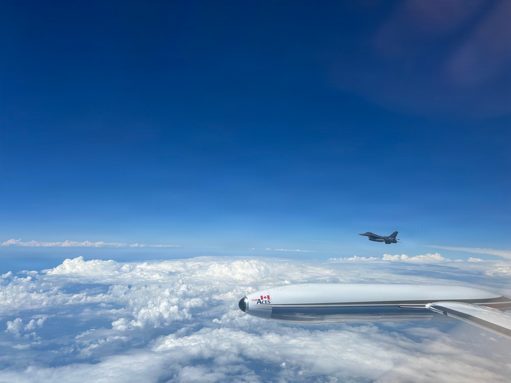 An aerial photograph taken from inside or alongside an aircraft showing a fighter jet flying in formation at high altitude. The foreground features a white engine nacelle or pod marked with 'TOP ACES' branding and a Canadian flag logo. A second aircraft, appearing to be an F-16 or similar fighter jet, is visible in the mid-distance against a deep blue sky above a thick cloud layer.