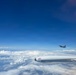 An aerial photograph taken from inside or alongside an aircraft showing a fighter jet flying in formation at high altitude. The foreground features a white engine nacelle or pod marked with 'TOP ACES' branding and a Canadian flag logo. A second aircraft, appearing to be an F-16 or similar fighter jet, is visible in the mid-distance against a deep blue sky above a thick cloud layer.
