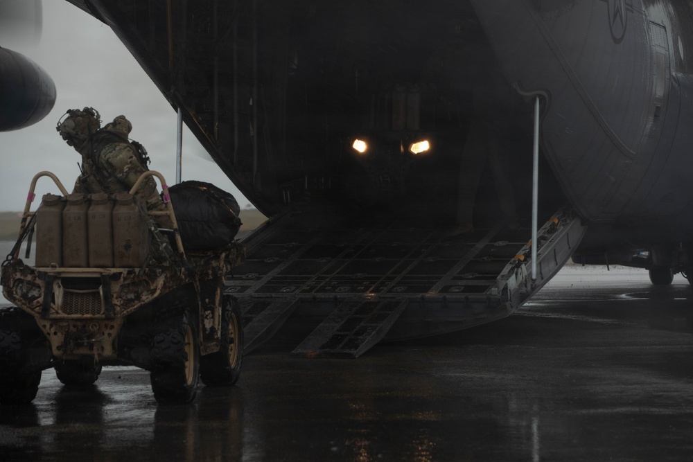 A fully-equipped soldier is riding an all-terrain vehicle (ATV) loaded with gear up the rear cargo ramp of a large military transport aircraft, likely a C-130 Hercules. The scene takes place on a wet tarmac under overcast, rainy conditions. Interior cargo bay lights illuminate the darkened aircraft interior.