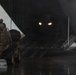 A fully-equipped soldier is riding an all-terrain vehicle (ATV) loaded with gear up the rear cargo ramp of a large military transport aircraft, likely a C-130 Hercules. The scene takes place on a wet tarmac under overcast, rainy conditions. Interior cargo bay lights illuminate the darkened aircraft interior.