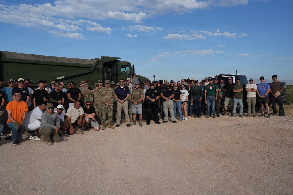 A large group of approximately 50-60 individuals poses for a group photograph in an arid, desert-like outdoor setting. The group includes military personnel in camouflage uniforms, law enforcement officers, and civilians in casual attire. Two large military-style vehicles are visible in the background, suggesting this may be a joint training exercise, community event, or operational briefing.
