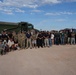A large group of approximately 50-60 individuals poses for a group photograph in an arid, desert-like outdoor setting. The group includes military personnel in camouflage uniforms, law enforcement officers, and civilians in casual attire. Two large military-style vehicles are visible in the background, suggesting this may be a joint training exercise, community event, or operational briefing.