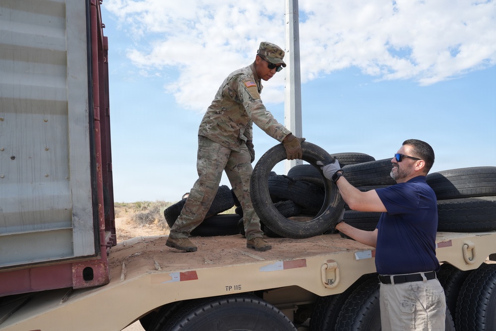 A U.S. Army soldier in OCP camouflage uniform and a civilian in a navy shirt are collaborating to load or unload used tires onto a flatbed military trailer marked 'TP 115'. The activity appears to take place in an arid, desert-like environment under a partly cloudy sky. The operation likely involves tire collection, disposal, or redistribution efforts.