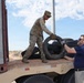A U.S. Army soldier in OCP camouflage uniform and a civilian in a navy shirt are collaborating to load or unload used tires onto a flatbed military trailer marked 'TP 115'. The activity appears to take place in an arid, desert-like environment under a partly cloudy sky. The operation likely involves tire collection, disposal, or redistribution efforts.
