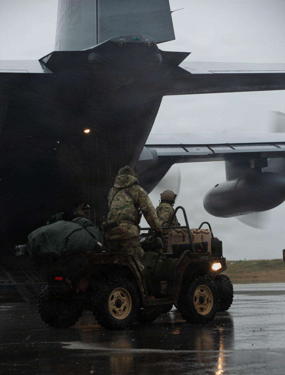 Military personnel in camouflage uniforms are loading or unloading a tactical all-terrain vehicle (ATV) onto or off a large military transport aircraft, likely a C-130 Hercules, via its rear cargo ramp. The operation is taking place on a wet tarmac in overcast, rainy conditions. At least two to three personnel are visible on or near the ATV, which has its headlights on.