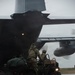 Military personnel in camouflage uniforms are loading or unloading a tactical all-terrain vehicle (ATV) onto or off a large military transport aircraft, likely a C-130 Hercules, via its rear cargo ramp. The operation is taking place on a wet tarmac in overcast, rainy conditions. At least two to three personnel are visible on or near the ATV, which has its headlights on.