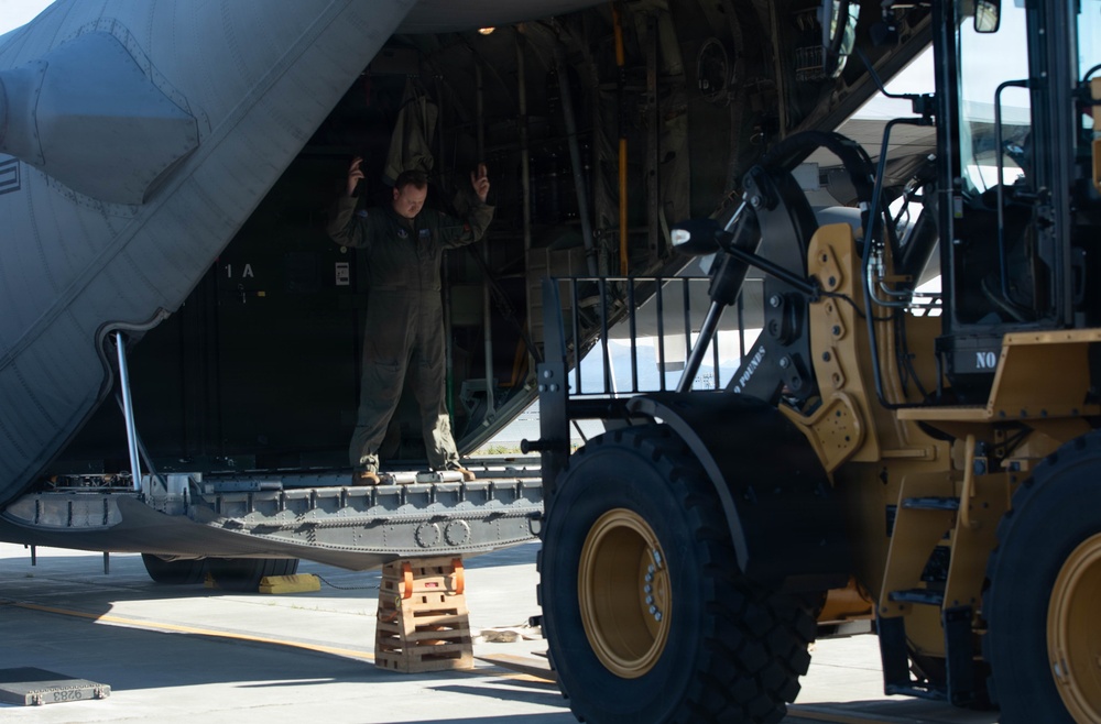 A military loadmaster or crew member in flight suit is directing a wheeled front-end loader (likely a Caterpillar model) toward the open rear cargo ramp of a large military transport aircraft, possibly a C-130 or C-17. The person is using hand signals to guide the vehicle during a loading or unloading operation on a tarmac. Wooden cribbing blocks are visible beneath the lowered cargo ramp for support.
