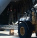 A military loadmaster or crew member in flight suit is directing a wheeled front-end loader (likely a Caterpillar model) toward the open rear cargo ramp of a large military transport aircraft, possibly a C-130 or C-17. The person is using hand signals to guide the vehicle during a loading or unloading operation on a tarmac. Wooden cribbing blocks are visible beneath the lowered cargo ramp for support.