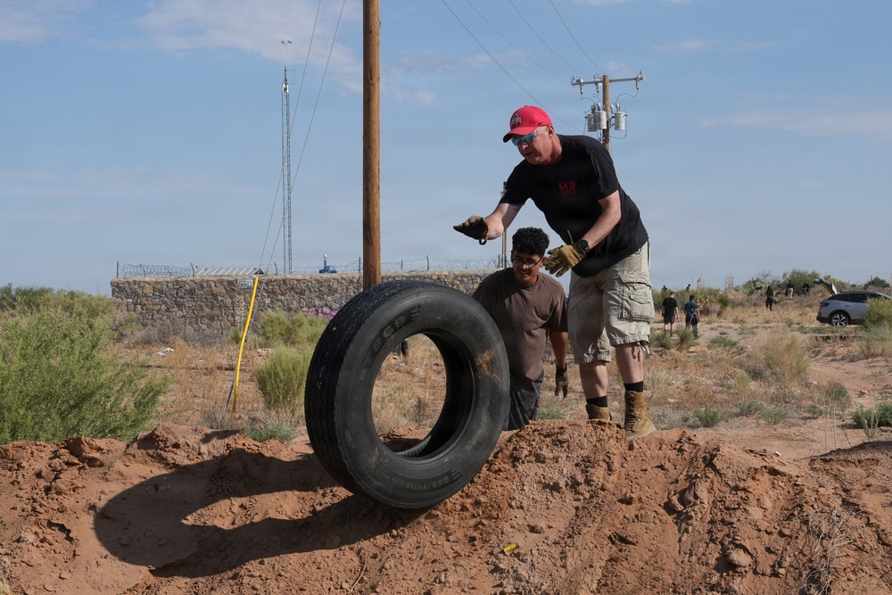 Two individuals are working together to move or position a large truck tire in a desert/arid environment. A man wearing a red cap, black t-shirt, and cargo shorts is guiding the tire while a younger person assists from behind. The setting appears to be a community cleanup or construction project in a dry, scrubland area.